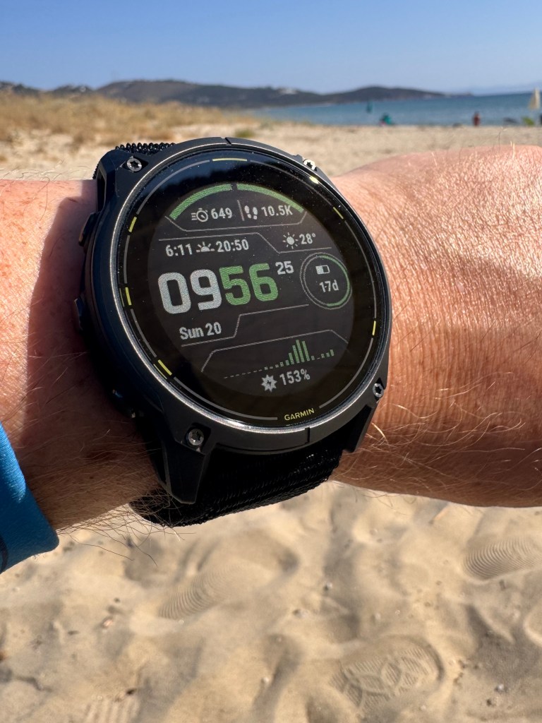 Close-up of a Garmin smartwatch displaying metrics such as steps taken, heart rate, and time, worn on a person's wrist against a sandy beach background.