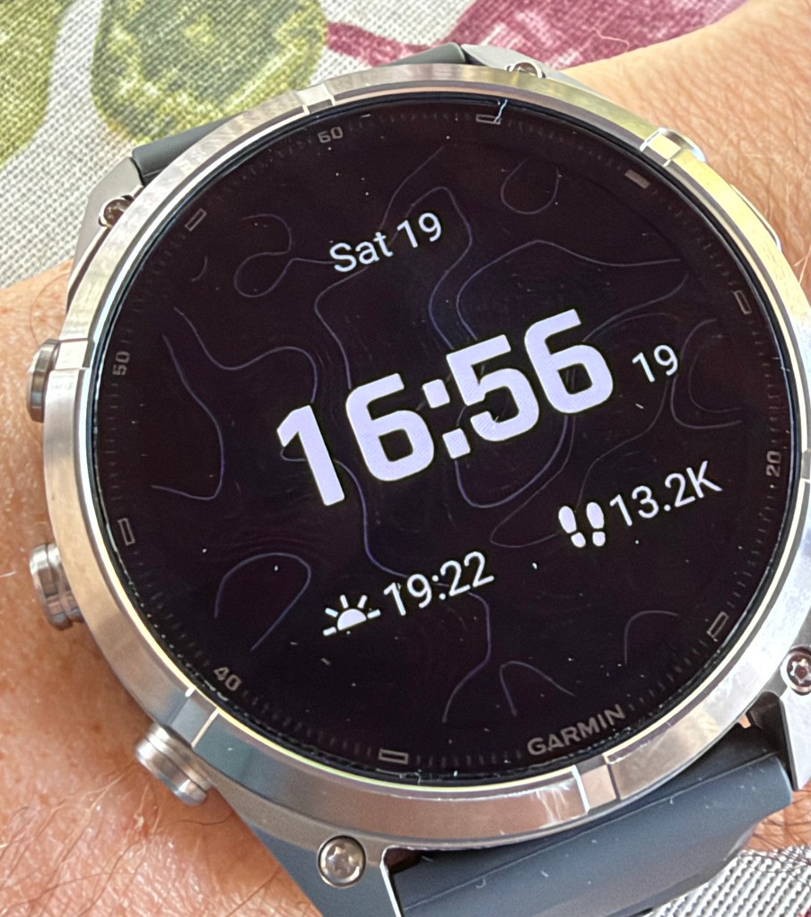 A close-up of a Garmin smartwatch displaying the time as 16:56 on Saturday the 19th, along with additional information about sunset time and step count.